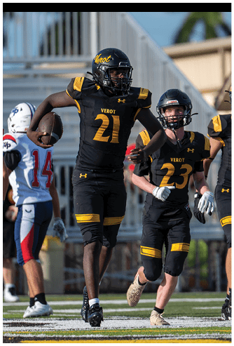Two people in black football uniforms with yellow accents are running on a field, one holding a football, while a person in a white and blue uniform stands nearby. Descripci n generada con IA