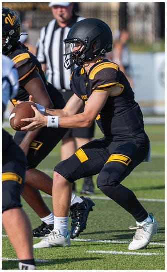 Two people in football uniforms and helmets are positioned closely, with one person holding a football and the other preparing to receive it. Descripci n generada con IA