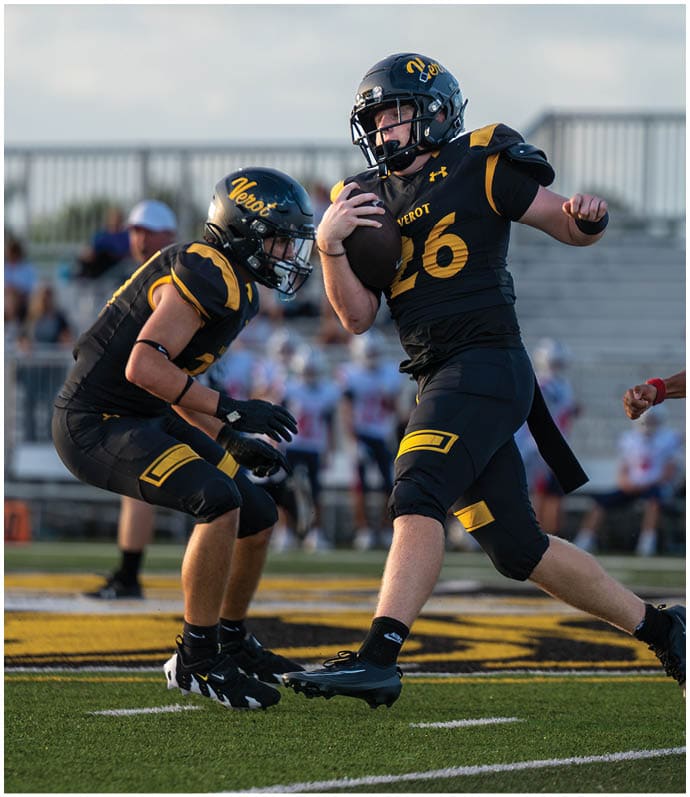 A person in black football gear is running with the ball while another person in black prepares to block and a person in white football gear lunges to tackle on a football field. Descripci n generada con IA