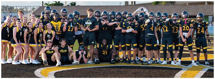 A group of people in football uniforms and cheerleading outfits pose together on a football field with a building in the background. Descripci n generada con IA
