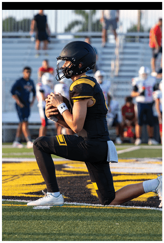 A person in a black and yellow football uniform kneels on one knee on a field while holding a football, with blurred people and bleachers in the background. Descripci n generada con IA