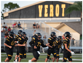 A group of people wearing black and yellow football uniforms are running on a field with a building in the background displaying the word VEROT. Descripci n generada con IA