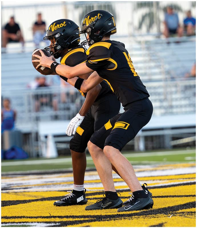 Two people in black and yellow football uniforms and helmets are positioned on a field with one holding a football ready to pass while the other prepares to receive it. Descripci n generada con IA