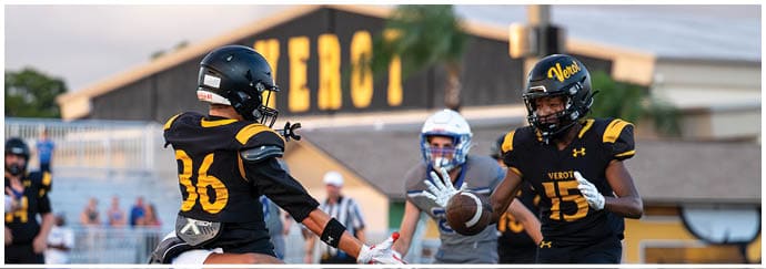 Two people in black and yellow football uniforms are playing on a field with one holding a football and the other reaching out to catch it while a referee and spectators are visible in the background. Descripci n generada con IA