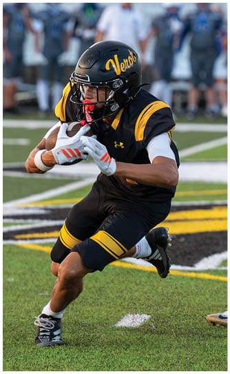 A person in a black and yellow football uniform is running with a football while another person reaches out with a gloved hand on a green field. Descripci n generada con IA