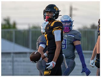 A person in a black and yellow football uniform holds a football on a field with other players in gray uniforms in the background. Descripci n generada con IA