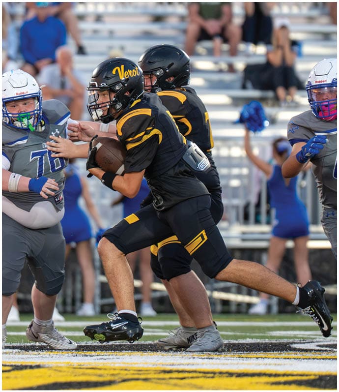Two people in black and yellow football uniforms are running with the ball while two people in gray and white uniforms attempt to block them on a football field. Descripci n generada con IA