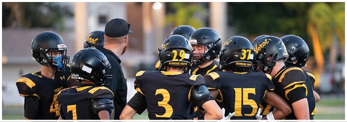 A group of people wearing black and yellow football uniforms and helmets are gathered closely together, seemingly in discussion or strategizing on a field. Descripci n generada con IA