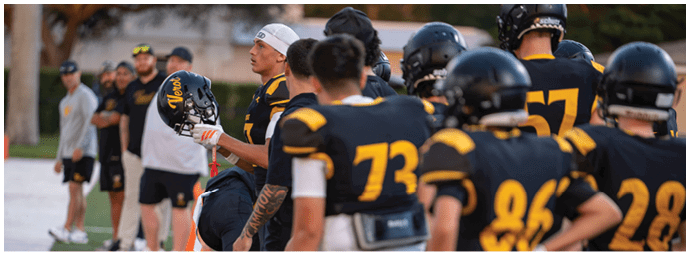 A group of people wearing black and yellow sports uniforms are gathered on a field, with some standing and others sitting, while a few people in casual clothing stand in the background. Descripci n generada con IA