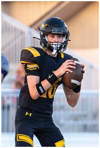A person wearing a black and yellow football uniform is holding a football in a ready-to-throw position on a grassy field with a referee standing nearby. Descripci n generada con IA
