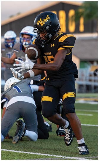 A person in a black and yellow football uniform catches a football while another person in a gray and white uniform kneels on the field during a game. Descripci n generada con IA