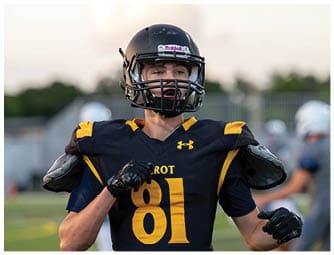 A person wearing a black and yellow football uniform and helmet is standing on a field with blurred players in the background. Descripci n generada con IA