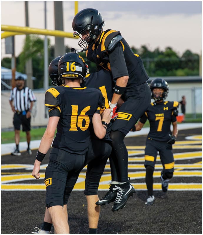 Two people wearing black and yellow football uniforms and helmets are engaged in a play on a football field near a yellow goalpost. Descripci n generada con IA