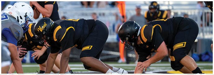 Two people in black and yellow football uniforms and helmets are crouched in a ready position on a football field. Descripci n generada con IA