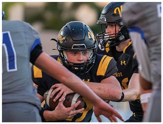 Two people wearing black and yellow football helmets and uniforms are focused and positioned closely during a game, with blurred opponents in gray uniforms in the foreground. Descripci n generada con IA