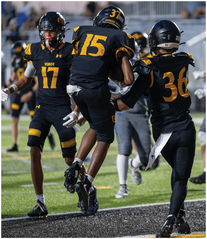 Three people in black and yellow football uniforms are on a field with one person jumping while holding a football and another reaching out to support them. Descripci n generada con IA