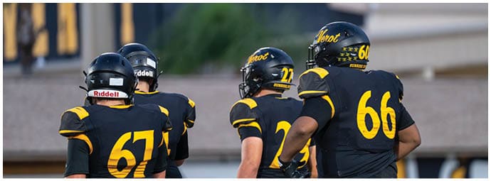 Four people wearing black and yellow football uniforms and helmets are standing on a field facing away from the camera. Descripci n generada con IA