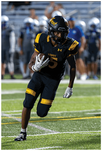 A person in a black and yellow football uniform is running on a field while holding a football, with other players visible in the background. Descripci n generada con IA