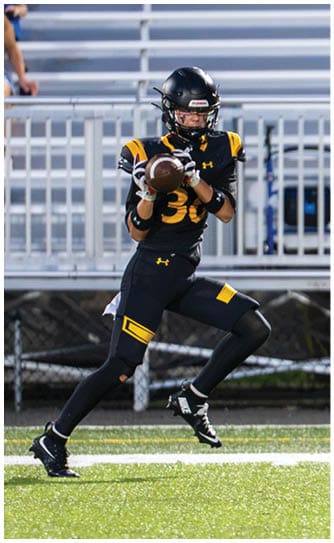 A person in black and yellow football gear is catching a football on a field with a metal bleacher background. Descripci n generada con IA