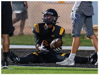 A person in a black and yellow football uniform is sitting on the ground with legs spread wide while holding a football, surrounded by two other people standing nearby on a sports field. Descripci n generada con IA