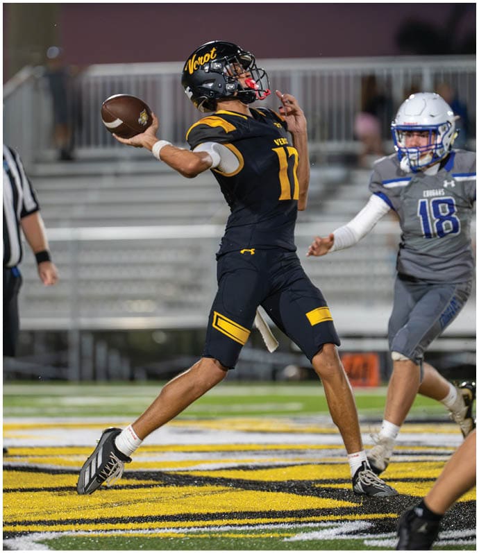 An adult in a black and yellow football uniform is throwing a football while another adult in a gray and blue uniform approaches on a football field. Descripci n generada con IA