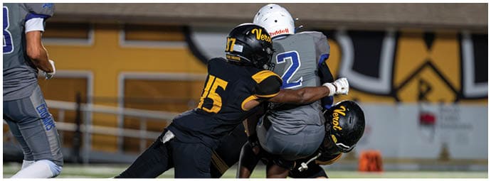 Two people in black and yellow football uniforms tackle a person in a gray and blue uniform on a football field while another person in gray and blue stands nearby. Descripci n generada con IA