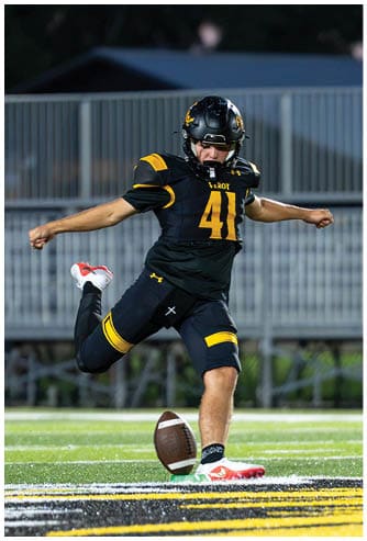 A person wearing a black and yellow football uniform is kicking a football on a field near the end zone. Descripci n generada con IA