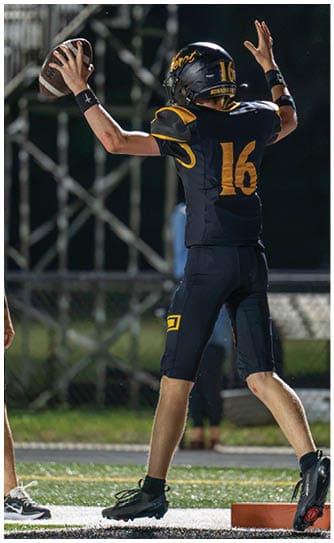 A person wearing a black and yellow football uniform with the number 16 is preparing to throw a football on a field near an orange marker. Descripci n generada con IA