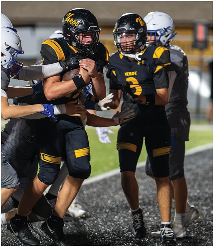 Two people in black and yellow football uniforms with helmets are on a field, with one holding a football tightly while being tackled by a person in a gray uniform and helmet. Descripci n generada con IA