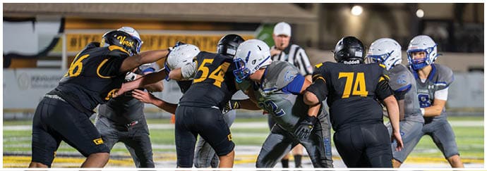People wearing football uniforms are engaged in a play on a lit outdoor field with a referee in the background. Descripci n generada con IA