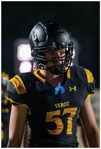 A person wearing a black and yellow football uniform with the number 57 and a helmet is standing on a field at night. Descripci n generada con IA