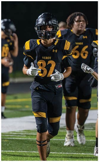 Two people in black and yellow football uniforms stand on a field at night, with one person wearing a helmet and gloves in the foreground. Descripci n generada con IA