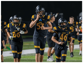Three people wearing black and yellow football uniforms with helmets are running on a field during a nighttime game or practice. Descripci n generada con IA
