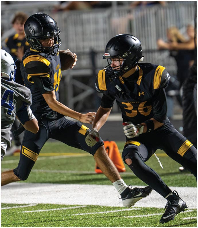 Three people in football uniforms are actively engaged in a play on a well-lit field with bleachers in the background. Descripci n generada con IA