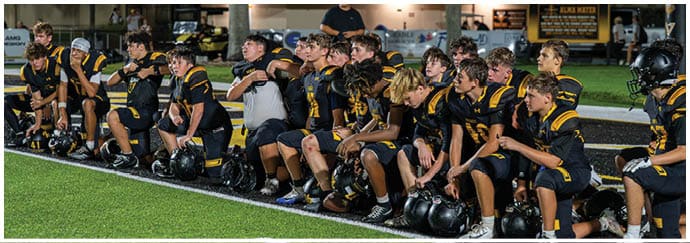 A group of people wearing black and yellow football uniforms sit on a bench on the sidelines of a football field. Descripci n generada con IA