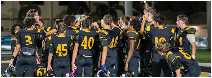A group of people wearing black and yellow football uniforms are gathered closely together on a field, appearing to be in a team huddle or discussion. Descripci n generada con IA