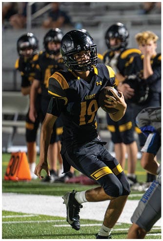 A person in a black and yellow football uniform is running with the ball while another person in a gray and blue uniform prepares to block on a football field. Descripci n generada con IA