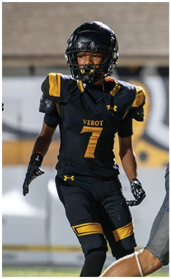 A person in a black and yellow football uniform with the number seven stands on a field while another person in gray sports gear is partially visible in the foreground. Descripci n generada con IA