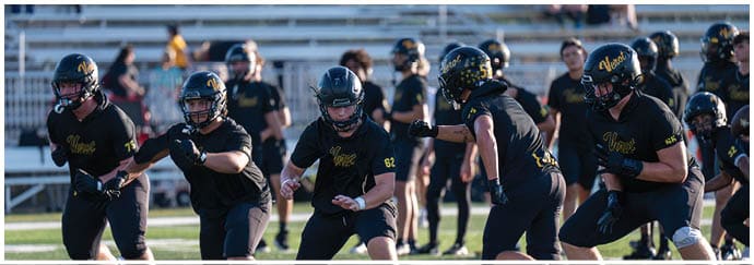 Five people in black sports uniforms and helmets are running on a football field during a game or practice. Descripci n generada con IA