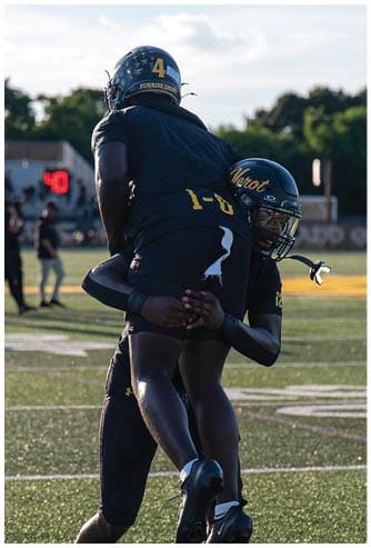 Two football players in black uniforms and helmets are celebrating on a field with one player lifting the other. Descripci n generada con IA