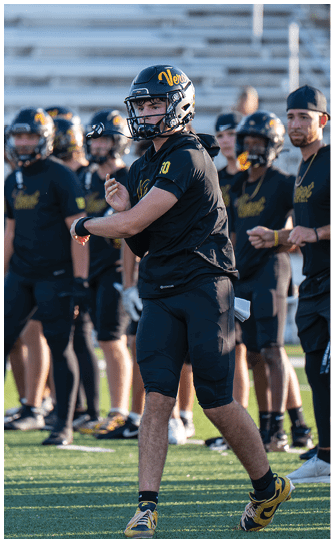 A group of people wearing black sports uniforms and helmets stand on a field with one person in the foreground preparing to throw a football. Descripci n generada con IA