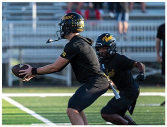 Two people wearing black sports uniforms and helmets with the word “Verot" are engaged in a football practice or game on a field with bleachers and spectators in the background. Descripci n generada con IA