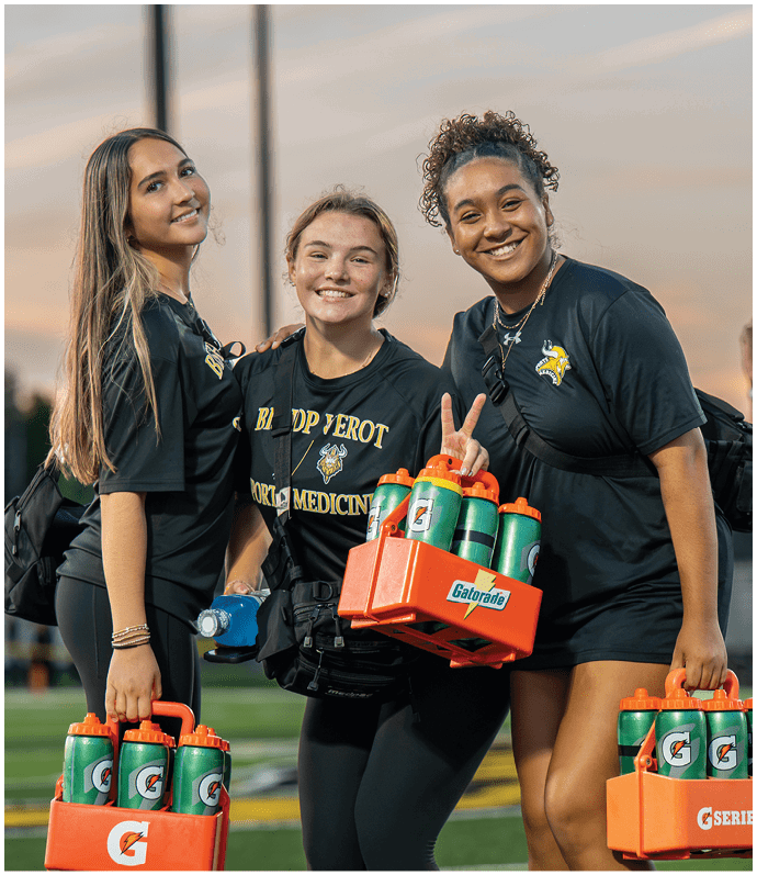 Three smiling people wearing matching sports shirts stand close together with one holding a carrier of water bottles and making a peace sign. Descripci n generada con IA