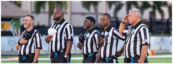 Five adults wearing black and white striped referee uniforms stand on a football field with their hands over their hearts. Descripci n generada con IA