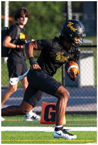 A person wearing a black football uniform and helmet is running on a green field while holding a football, with another person standing in the background. Descripci n generada con IA