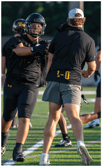 An adult wearing a black football uniform and helmet is reaching out toward another adult in a black shirt and gray shorts on a sports field. Descripci n generada con IA