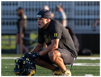 An adult wearing athletic clothing and a headband is crouched on a sports field holding a football helmet while looking intently to the side. Descripci n generada con IA