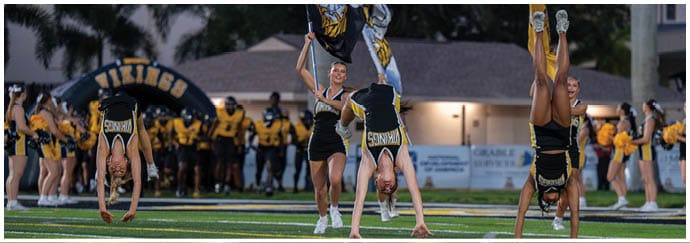 People in coordinated outfits perform a cheerleading routine with flags and high kicks in front of a crowd near a building and palm trees. Descripci n generada con IA