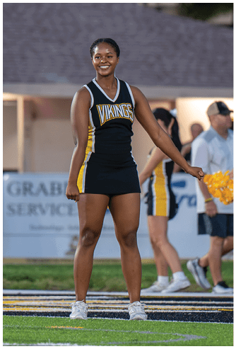 A person wearing a black and yellow cheerleading uniform with the word Vikings on the front is smiling and standing with one arm extended. Descripci n generada con IA