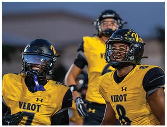 Two adults wearing yellow and black football uniforms with helmets are smiling and standing on a field with another player in the background. Descripci n generada con IA